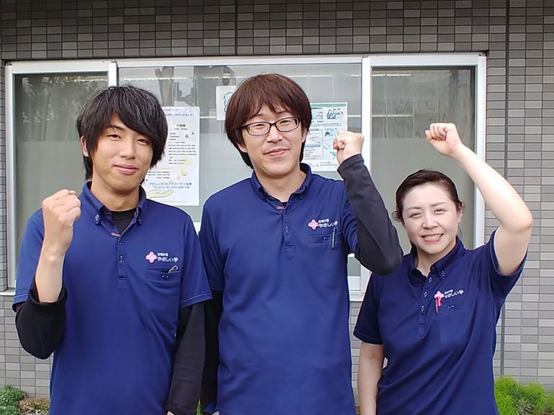 施設スタッフ3名が建物前で記念撮影。青いポロシャツに統一された制服を着用。