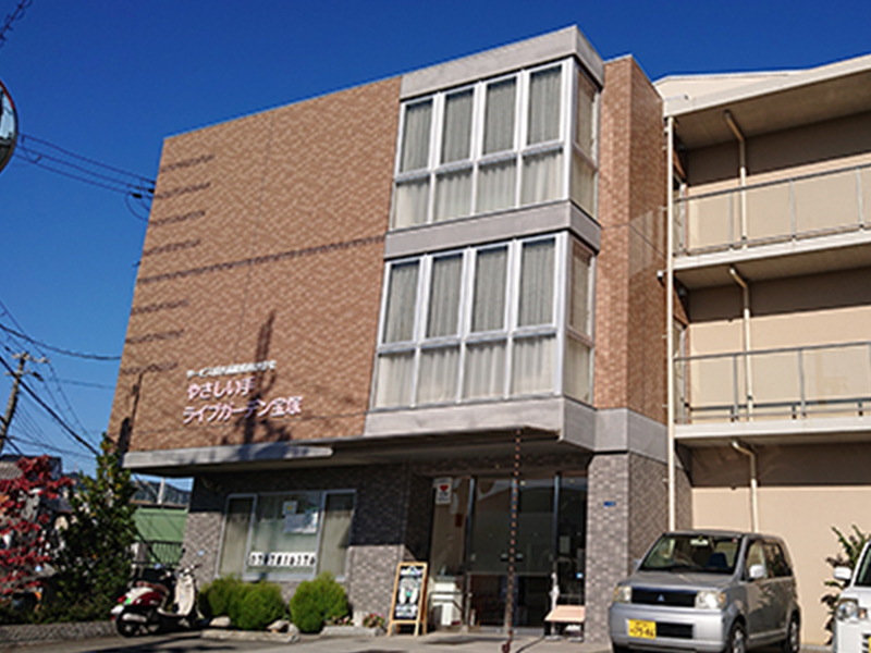 赤レンガ造りの介護施設の外観。晴天下の建物全景で、玄関と駐車スペースが見える。